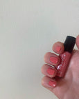 Hand holding a bottle of Rosy - Clean Nail Polish with nails painted in a matching rosy coral shade, highlighting clean beauty and vegan nail polish.