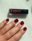 Hand with nails painted deep burgundy next to a bottle of Velvet - Clean Nail Polish by HOOPLA on a light surface, showcasing smooth, glossy finish.
