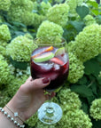 Hand holding a glass of sangria with fruit slices, showing nails painted in deep red using Sangria - Clean Nail Polish, set against a background of green hydrangea flowers.