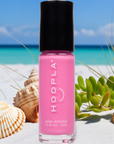 Bottle of Pink Flamingo - Clean Nail Polish with bright pink color standing on white sand next to seashells and green beach plants under a clear blue sky.