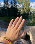 Hand with neatly manicured nails painted in soft, natural shade using Angelic Arrow - Clean Nail Polish, adorned with silver and pearl rings, against an outdoor background.