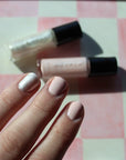 Close-up of a hand with soft nude and pearly nail polish on fingers, showcasing Angel - Clean Nail Polish bottles on a pink and white checkered background.