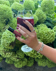 Hand with polished nails in deep red shade holding a glass of sangria and a bottle of Sangria - Clean Nail Polish, set against green foliage background.