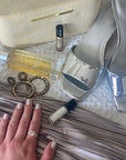 Hand with nails painted in soft neutral shade using Mini Pearl - Clean Nail Polish, surrounded by silver shoes, perfume, and accessories on a fabric surface.
