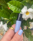 Hand holding a bottle of HOOPLA light lavender nail polish with matching painted nails, set against a background of green leaves and white flowers, featuring M+M - Clean Nail Polish.