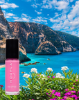 Pink Flamingo - Clean Nail Polish bottle with bright pink color displayed against a scenic coastal backdrop featuring turquoise ocean, rocky cliffs, and vibrant flowers.