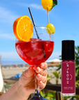 Hand holding a glass of red sangria garnished with an orange slice and a grape with a pineapple pick against a sunny beach background, next to a bottle of Sangria - Clean Nail Polish by HOOPLA.