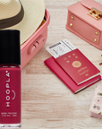 Sangria - Clean Nail Polish bottle in a rich red shade placed next to travel essentials including a pink suitcase, wide-brimmed hat, Japan passport, sunglasses, and a pink purse, highlighting a stylish clean beauty travel theme.
