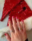 Hand with pink polished nails wearing silver rings resting on a red sequined fabric with a small bottle of Private School Girl - Clean Nail Polish nearby.