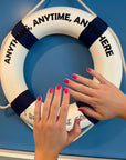 Hands with glossy pink nails polished using Rosy - Clean Nail Polish held up against a white and navy life preserver ring with the text "Anything, Anytime, Anywhere" on a blue background.