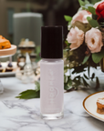 A bottle of Angel - Clean Nail Polish with a black cap displayed on a marble table, surrounded by flowers and plates with desserts in a cozy setting.