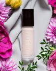Bottle of Fluff - Clean Nail Polish in a soft neutral shade surrounded by pink, purple, and yellow flowers on a light fabric background, highlighting a clean beauty nail treatment.