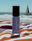 A bottle of Newport Love - Clean Nail Polish in a purple shade standing on a striped towel by the beach with the ocean and a sailboat in the background.