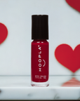 A small bottle of HOOPLA Velvet - Clean Nail Polish in deep red shade with black cap, placed on a reflective surface with red heart shapes blurred in the background.