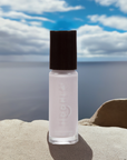 Bottle of Angel - Clean Nail Polish with a pale, neutral shade standing on a sandy surface under a cloudy sky by the water, highlighting clean beauty and nail care.