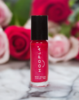 Small bottle of HOOPLA red nail polish labeled Private School Girl - Clean Nail Polish, displayed on a marble surface with blurred pink and red roses in the background
