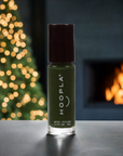 A bottle of HOOPLA vegan nail polish in a deep green shade, placed on a surface with a softly blurred Christmas tree and cozy fireplace in the background, showcasing Holly - Clean Nail Polish.