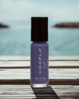 Bottle of Newport Love - Clean Nail Polish in a muted lavender shade from Hoopla, standing on a wooden surface with a blurred ocean background.