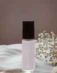 A bottle of Angel - Clean Nail Polish with a soft pink shade, placed next to delicate white dried flowers on a neutral background, highlighting clean beauty and vegan nail care.