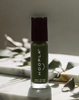 Holly - Clean Nail Polish in a dark green shade by HOOPLA, shown in a small bottle with black cap on a sunlit surface with green leaves nearby, representing clean beauty nail polish.