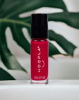 HOOPLA Pocket Nail Polish - Blissful - vegan, cruelty free, made in the USA, vibrant red mini nail polish bottle with black cap on white surface and green leafy background.