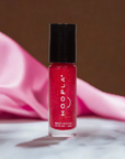 A bottle of Pink Foil - Clean Nail Polish by HOOPLA featuring a vibrant pink foil shimmer with a black cap, set on a marble surface with soft pink fabric in the background.