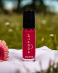 Bottle of HOOPLA Pocket Nail Polish - Blissful - vegan, cruelty free, made in the USA, shown in vibrant red with a black cap, placed on a soft surface next to a pink flower in an outdoor setting.