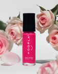 Bright pink nail polish bottle with black cap labeled HOOPLA, placed among soft pink roses and rose petals, showcasing the Rosy - Clean Nail Polish in a clean beauty setting.