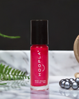 A vibrant red bottle of Private School Girl - Clean Nail Polish by HOOPLA, standing on a reflective marble surface with green fern leaves and a string of gray pearls nearby.