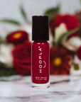 A small bottle of Hoopla nail polish in a deep red shade with black cap, displayed against a blurred background of red and white flowers, featuring Velvet - Clean Nail Polish.