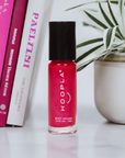 A small bottle of hoopla nail polish in a vibrant red shade standing on a white surface next to pink and white books and a green potted plant, labeled Private School Girl - Clean Nail Polish.