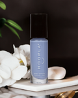 A bottle of M+M - Clean Nail Polish in a soft lavender shade placed on a white surface with white flowers and green leaves in the background, showcasing vegan clean beauty.
