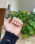 Close-up of a hand with neatly manicured nails painted in soft white, showcasing Angelic Arrow - Clean Nail Polish against a backdrop of green leafy plants with orange flowers.