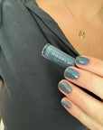 Hand holding a bottle of HOOPLA nail polish with nails painted in muted blue-gray shade, wearing a dark gray top and a gold heart-shaped necklace, featuring Diesel - Clean Nail Polish.