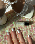 Hand with nails painted in shiny pearl color using Mini Pearl - Clean Nail Polish, placed near a pair of white sandals adorned with pearl decorations on a sequined surface.