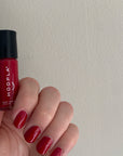 Hand holding a bottle of HOOPLA Pocket Nail Polish - Blissful - vegan cruelty free made in the USA with nails painted in a shiny deep red shade against a plain light background