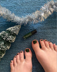 Close-up of feet with dark green nail polish painted on toenails next to a small snowy pine tree, silver tinsel, and a bottle of Holly - Clean Nail Polish on a blue carpet.