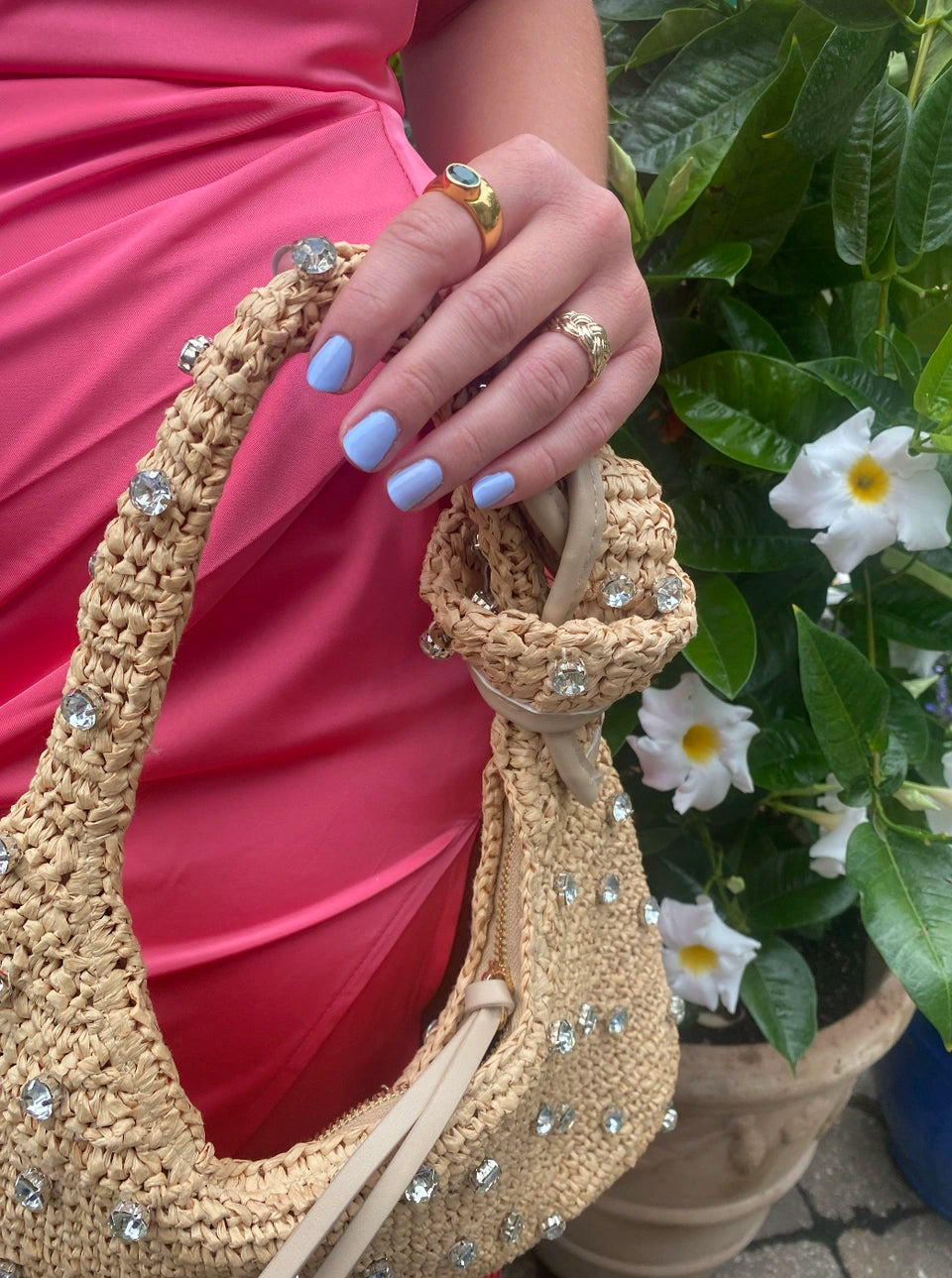 Hand with light blue nails painted using M+M - Clean Nail Polish holding a woven beige bag adorned with crystal decorations, dressed in vibrant pink fabric, with green foliage and white flowers in the background.