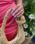 Hand with light blue nails painted using M+M - Clean Nail Polish holding a woven beige bag adorned with crystal decorations, dressed in vibrant pink fabric, with green foliage and white flowers in the background.