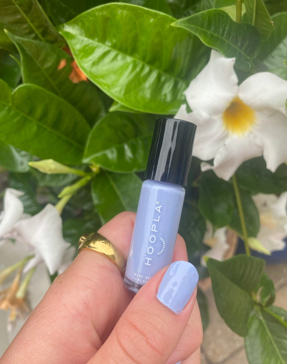 Hand holding a bottle of HOOPLA light lavender nail polish with matching painted nails, set against a background of green leaves and white flowers, featuring M+M - Clean Nail Polish.