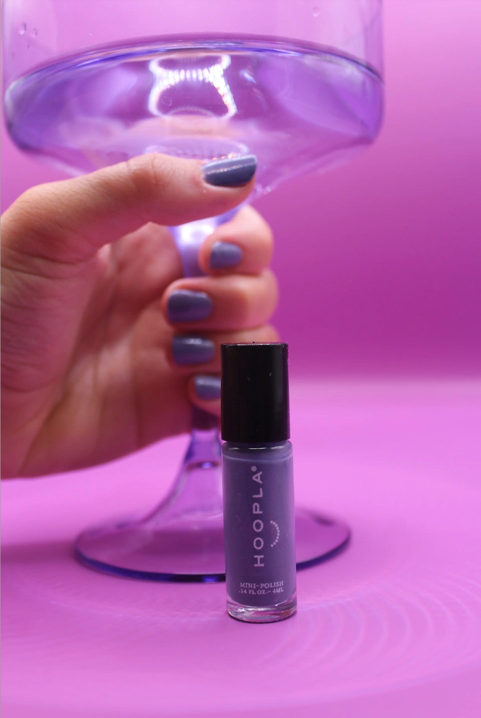 Hand with nails painted in muted purple holding a transparent purple stemmed glass above a bottle of Newport Love - Clean Nail Polish on a soft purple background