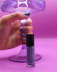 Hand with nails painted in muted purple holding a transparent purple stemmed glass above a bottle of Newport Love - Clean Nail Polish on a soft purple background