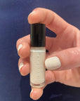 Hand holding a small bottle of Fluff - Clean Nail Polish with matching clean white nail polish on nails against a dark blue background.
