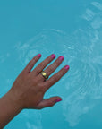 Hand with nails painted bright pink using Pink Flamingo - Clean Nail Polish, submerged partially in clear blue water, showcasing vibrant clean beauty nail treatment.