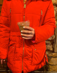 Person wearing a bright orange jacket holding a drink with nails painted in white using Angel - Clean Nail Polish, showcasing a clean beauty vegan nail polish look.