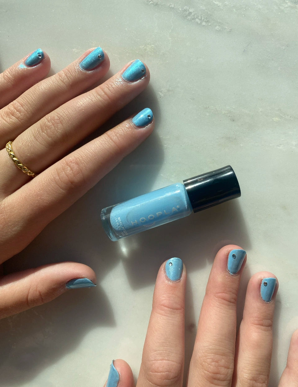 Hands with blue nail polish decorated with small black dots next to a bottle of Sky - Clean Nail Polish on a light surface, showcasing a clean beauty vegan nail treatment.