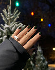 Hand with dark green nail polish applied, shown outdoors at night near snowy pine trees and colorful holiday lights, featuring "Holly - Clean Nail Polish" on the nails.