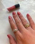 Close-up of a hand with nails painted in rosy pink using Rosy - Clean Nail Polish, featuring gold and silver knot rings on the fingers and the nail polish bottle in the background on a marble surface.