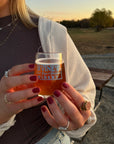 Hands with dark red nail polish holding a glass of amber liquid outdoors at sunset, showcasing Velvet - Clean Nail Polish in a cozy casual setting.