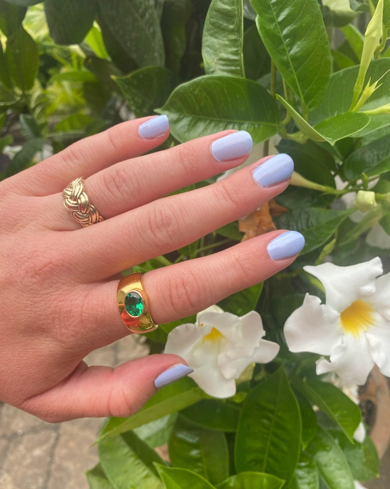 Close-up of a hand with light lavender nails painted with M+M - Clean Nail Polish, adorned with gold rings, gently resting near white flowers and green leaves outdoors.